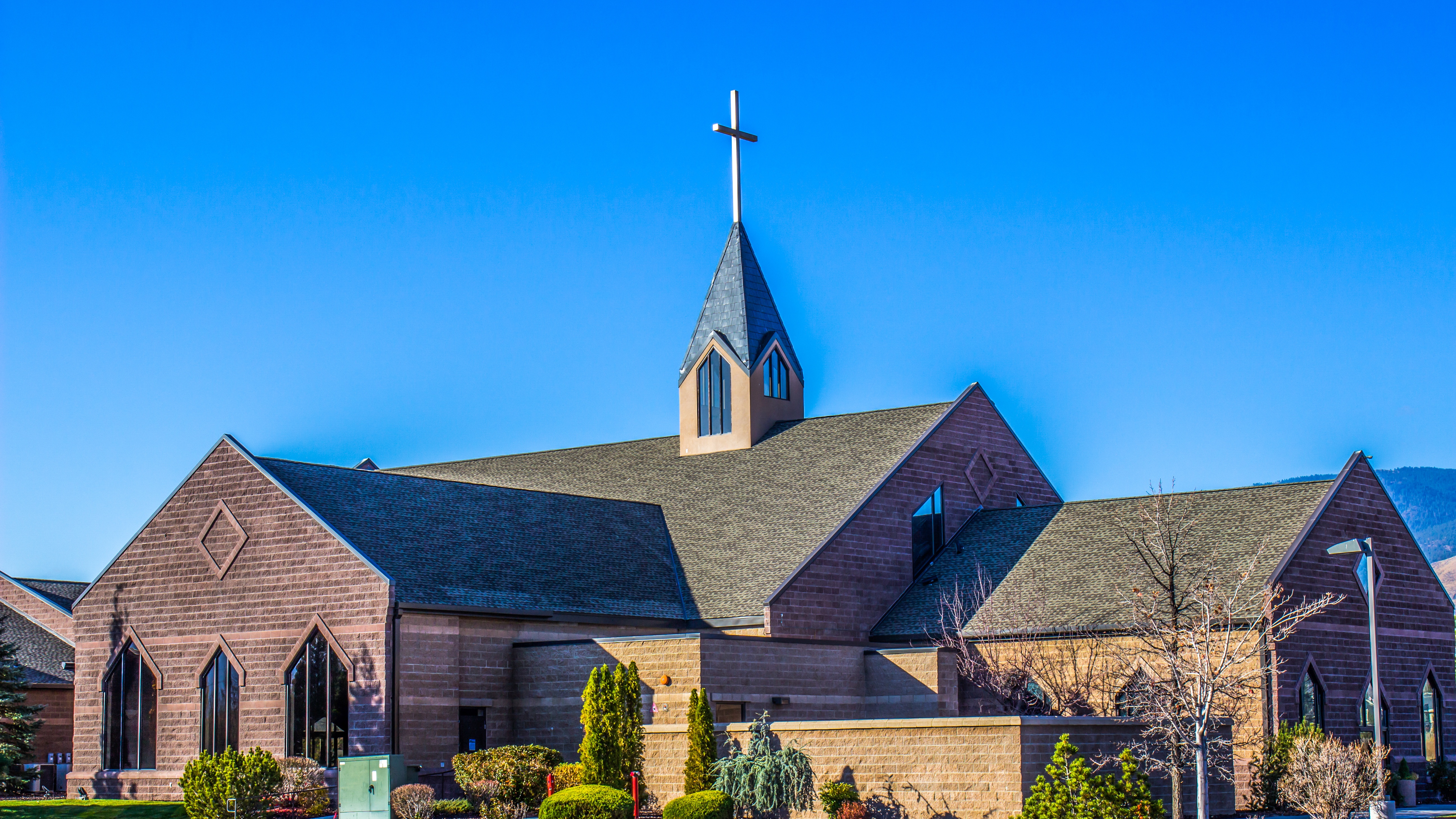 A large church with a steeple and cross, gray asphalt shingle roofing, and blue sky in the background