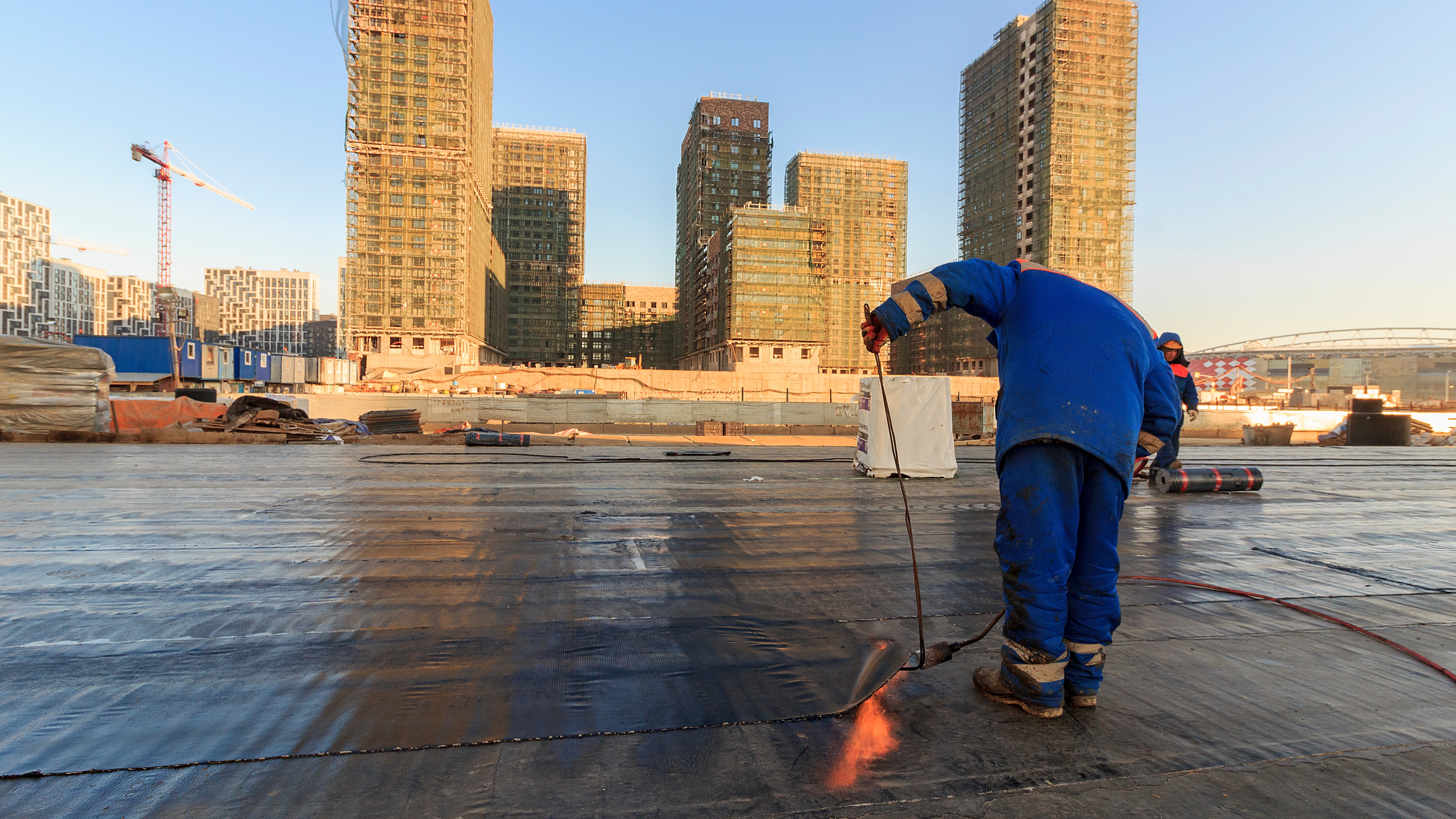 A commercial roofing restoration contractor applying a waterproof membrane to a flat roof with large multi-story buildings in the background